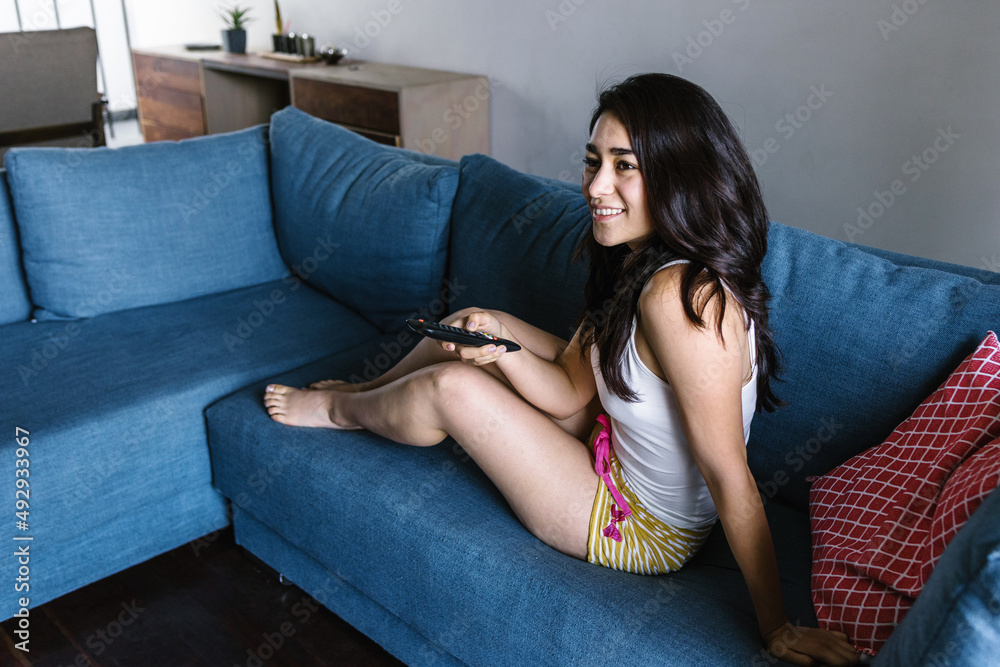 young latin woman lying relaxing on sofa in living room and watching TV at home in Mexico Latin America young latin woman lying relaxing on sofa in living room and watching TV at home in Mexico Latin America