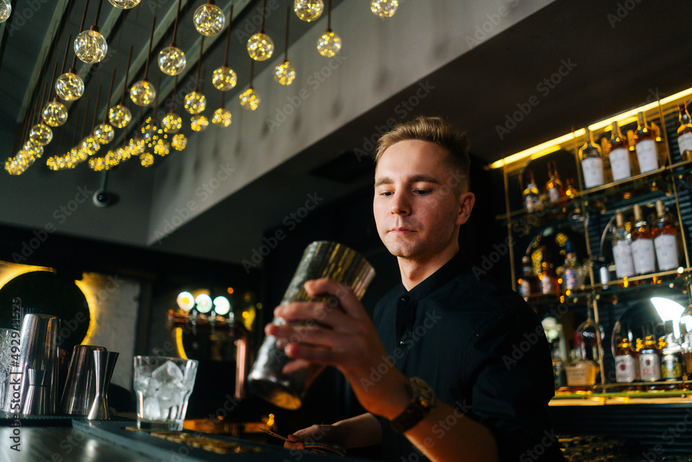 Close-up low-angle view of young professional barman making refreshing alcoholic cocktail standing behind bar counter in modern dark nightclub, on background of shelves with different alcoholic drinks