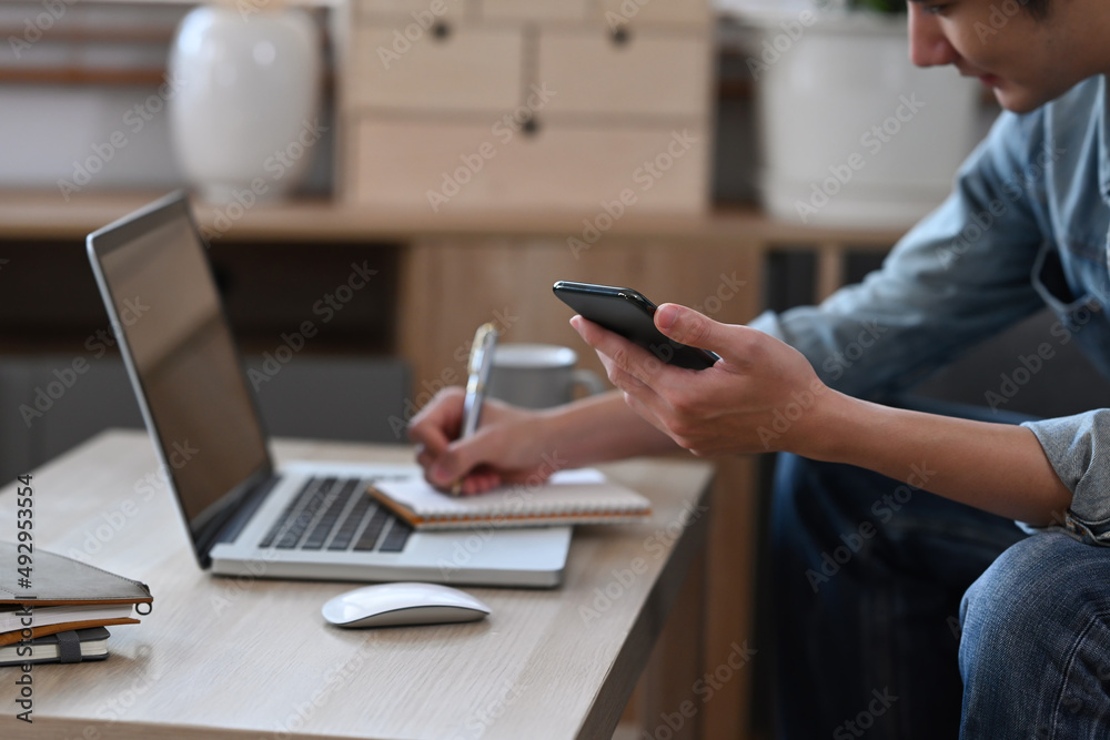 Fototapeta premium Young man holding smart phone and writing ideas in a notebook.