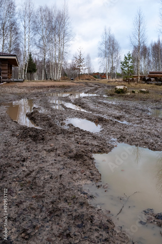 Rural muddy dirt road late autumn after rain. Off-road, wild dirt trail with large puddles background village, showing difficulties movement people in rural areas during rainy season. Vertical photo