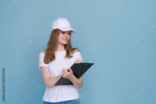 Delivery woman in white uniform isolated on blue background. Girl in white cap and t-shirt, blue jeans, working as a courier or dealer, holding a folder with papers writes. Copy space