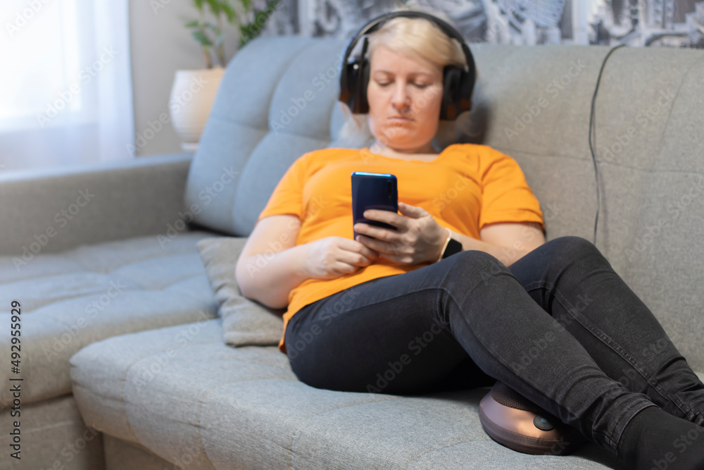 A girl in headphones and with a phone in her hands, sitting on the couch, makes a foot massage with a massage pillow. A girl in headphones and with a phone in her hands, sitting on the couch, makes a foot massage with a massage pillow.