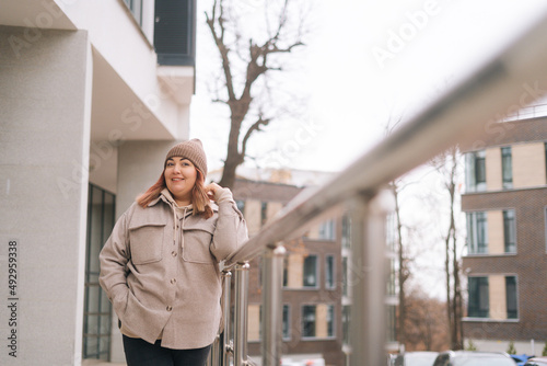 Portrait of happy overweight woman in warm hat and jacket standing near railing of office building at city street in cloudy autumn day, looking at camera. Freezing female waiting for someone outdoors.