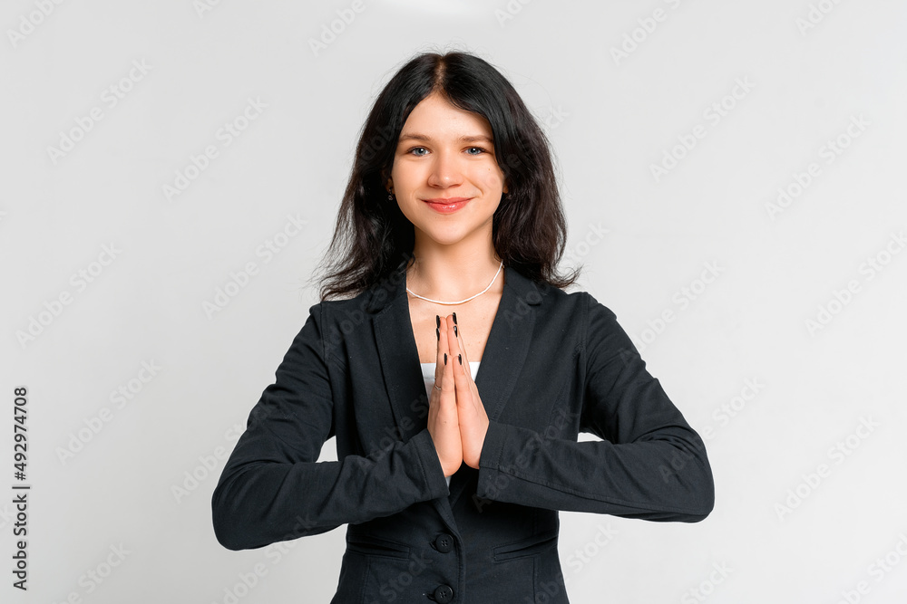 Portrait of calm young dark-haired girl praying, clasp hands together and making wish, pleading, standing in black blazer over grey background Portrait of calm young dark-haired girl praying, clasp hands together and making wish, pleading, standing in black blazer over grey background