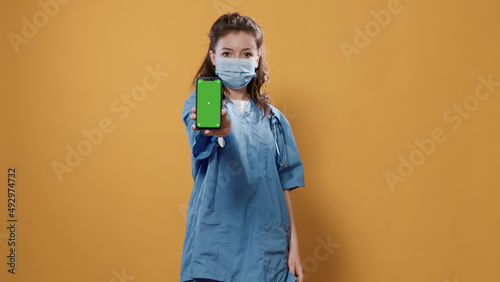 Doctor with stethoscope holding smartphone and texting showing green screen mockup at camera. Medic in hospital uniform wearing surgical mask using touchscreen device presenting copy space.