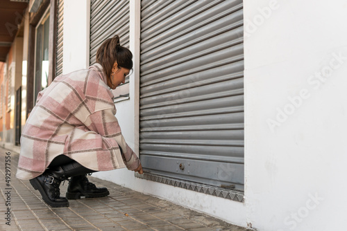 A close up shot of a young woman lowering the metal fence of her business at the time of closure. Brunette girl crouching to close her sales room once her working day is over.