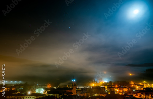Cityscape in the night with stars in the sky. City in mist. Europe building in mist. City and orange light in the night. City covered with fog. Foggy sky. Old town in Europe. Picturesque cityscape.