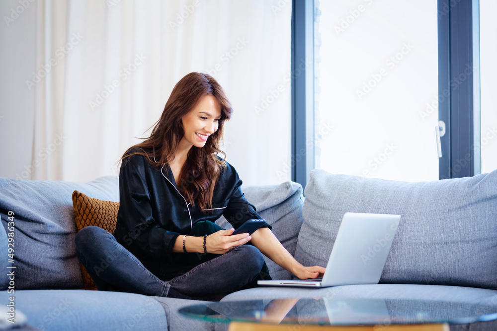 Attractive brunette haired woman using her mobile phone and laptop while working from home Attractive brunette haired woman using her mobile phone and laptop while working from home