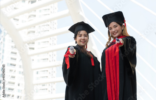 Two Asian young beautiful graduate female students with master and bachelor degree standing smiling and holding diploma in hand expressing proud face. Blur background of University building