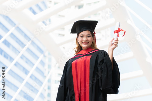 Happy Asian young beautiful graduate female student with University degree standing and holding diploma in raised hand after graduation wearing black cap with red tassels. Blur background of building