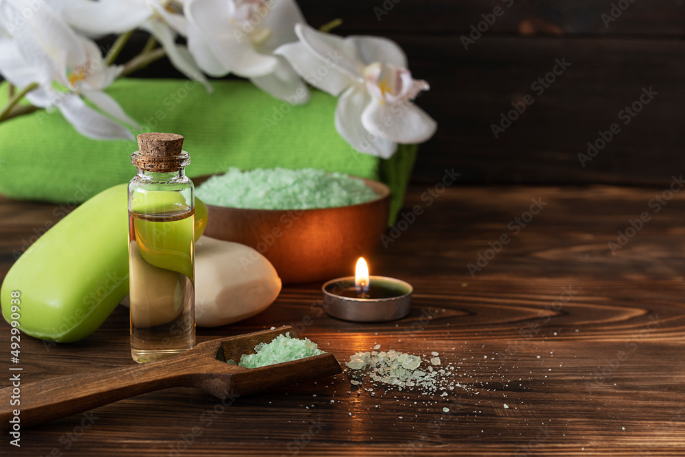 Body oil in a clear bottle, fragrant soap and bath salt on a dark table. Green bath towel and orchid sprig in the background Body oil in a clear bottle, fragrant soap and bath salt on a dark table. Green bath towel and orchid sprig in the background