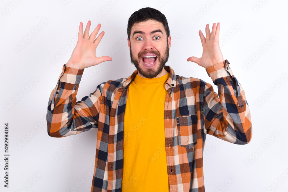 young caucasian man wearing plaid shirt over white background looks with excitement at camera, keeps hands raised over head, notices something unexpected, reacts on sudden news. young caucasian man wearing plaid shirt over white background looks with excitement at camera, keeps hands raised over head, notices something unexpected, reacts on sudden news.