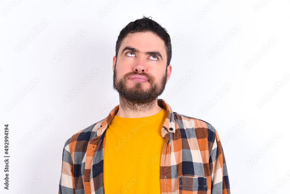 young caucasian man wearing plaid shirt over white background has worried face looking up lips together, being upset thinking about something important, keeps hands down. young caucasian man wearing plaid shirt over white background has worried face looking up lips together, being upset thinking about something important, keeps hands down.