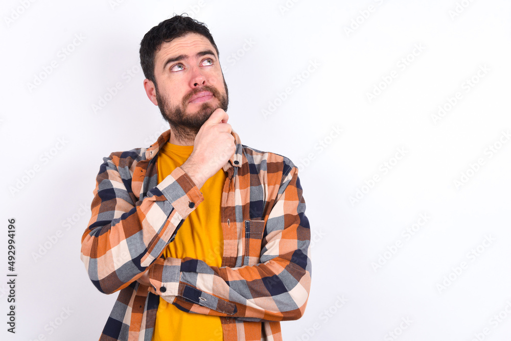Portrait of thoughtful young caucasian man wearing plaid shirt over white background keeps hand under chin, looks away trying to remember something or listens something with interest. Youth concept. Portrait of thoughtful young caucasian man wearing plaid shirt over white background keeps hand under chin, looks away trying to remember something or listens something with interest. Youth concept.