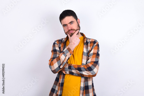 Thoughtful smiling young caucasian man wearing plaid shirt over white background keeps hand under chin, looks directly at camera, listens something with interest. Youth concept.