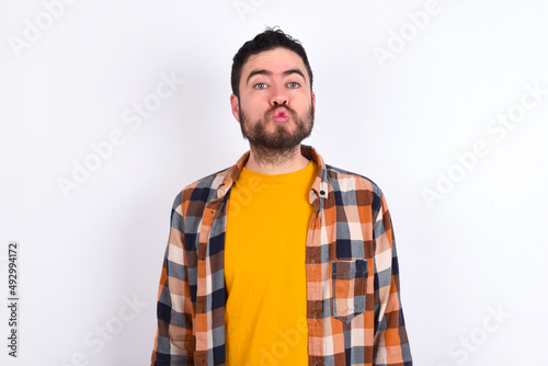 young caucasian man wearing plaid shirt over white background, keeps lips as going to kiss someone, has glad expression, grimace face. Standing indoors. Beauty concept.