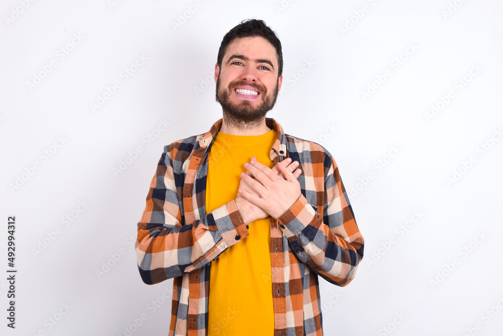 Honest young caucasian man wearing plaid shirt over white background keeps hands on chest, touched by compliment or makes promise, looks at camera with great pleasure. Honest young caucasian man wearing plaid shirt over white background keeps hands on chest, touched by compliment or makes promise, looks at camera with great pleasure.