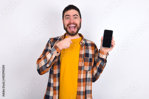 Excited young caucasian man wearing plaid shirt over white background holding and pointing with finger at smartphone with blank screen. Advertisement concept. © Jihan