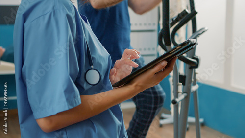 Health specialist in uniform using digital tablet to help senior patient with physical injury and mechanical disorders. Nurse holding device at muscle rehabilitation procedure. Close up.