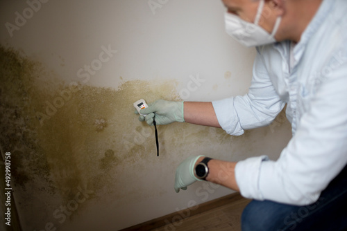Man with nose mouth protection measures the moisture level on a wall with mold in an apartment