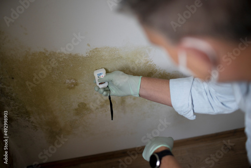 Man with nose mouth protection measures the moisture level on a wall with mold in an apartment