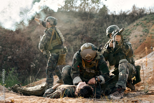 responsive army soldiers with injured comrade calling for help, blowing smoke, marines team in action, surrounded fire and smoke, shooting with assault rifle and machine gun, attacking enemy