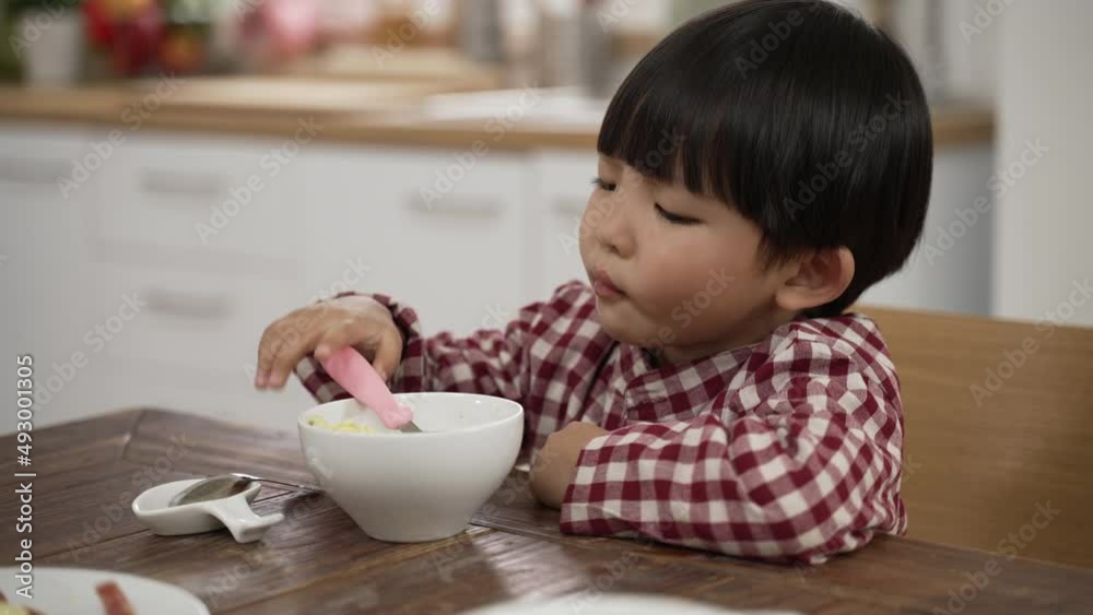 closeup of unhappy Asian little boy making a disgust expression while looking at green vegetable in bowl and shaking head show dislike at dining table