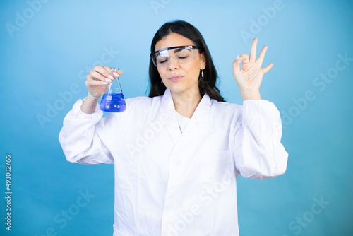 Young brunette woman wearing scientist uniform holding test tube over isolated blue background relax and smiling with eyes closed doing meditation gesture with fingers