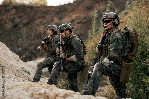 Team of armed special forces soldiers with rifles standing together looking at side, planning strategy of shooting, waiting for enemy. confident serious soldiers in military clothes outdoors