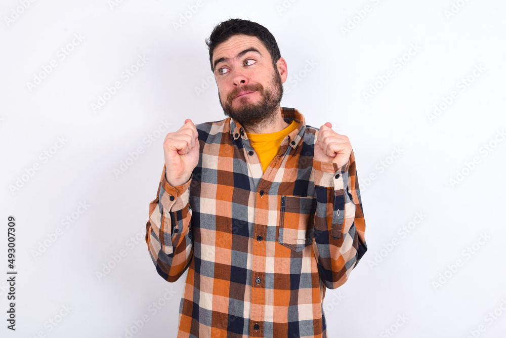 young caucasian man wearing plaid shirt over white background clenches fists and awaits for something nice happened looks away bites lips and waits announcement of results young caucasian man wearing plaid shirt over white background clenches fists and awaits for something nice happened looks away bites lips and waits announcement of results
