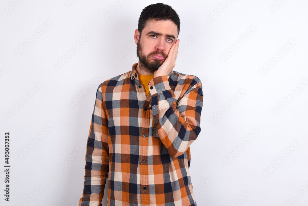 Sad lonely young caucasian man wearing plaid shirt over white background touches cheek with hand bites lower lip and gazes with displeasure. Bad emotions Sad lonely young caucasian man wearing plaid shirt over white background touches cheek with hand bites lower lip and gazes with displeasure. Bad emotions