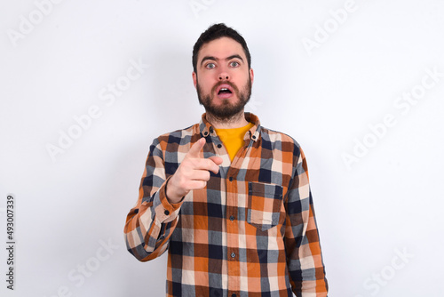 Shocked young caucasian man wearing plaid shirt over white background points at you with stunned expression