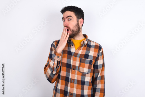 young caucasian man wearing plaid shirt over white background covers mouth and looks with wonder at camera, cannot believe unexpected rumors.