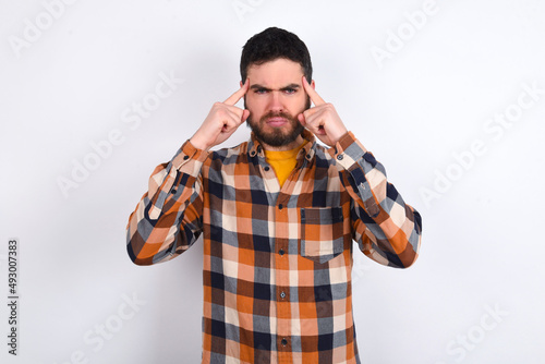 Serious concentrated young caucasian man wearing plaid shirt over white background keeps fingers on temples, tries to ease tension, gather with thoughts and remember important information for exam
