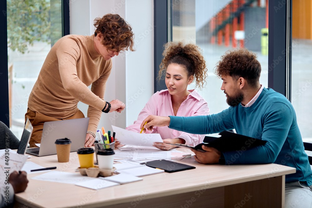 male employee discussing online project with coworkers, talking about presentation to skilled team leaders sitting behind desk. Friendly diverse colleagues share opinions, discuss