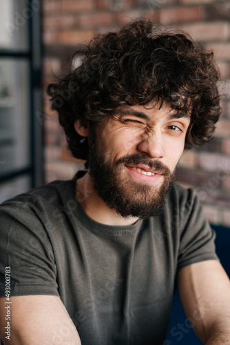 Close-up face of positive handsome young man winking looking at camera sitting on chair in cozy living room with brick wall. Closeup vertical shot of confident bearded curly male posing at home.