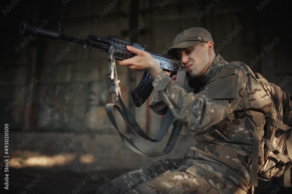 a young Ukrainian soldier in a gray camouflage uniform wearing a cap and tactical black glasses sits on a concrete floor inside a large destroyed building and aims into the distance