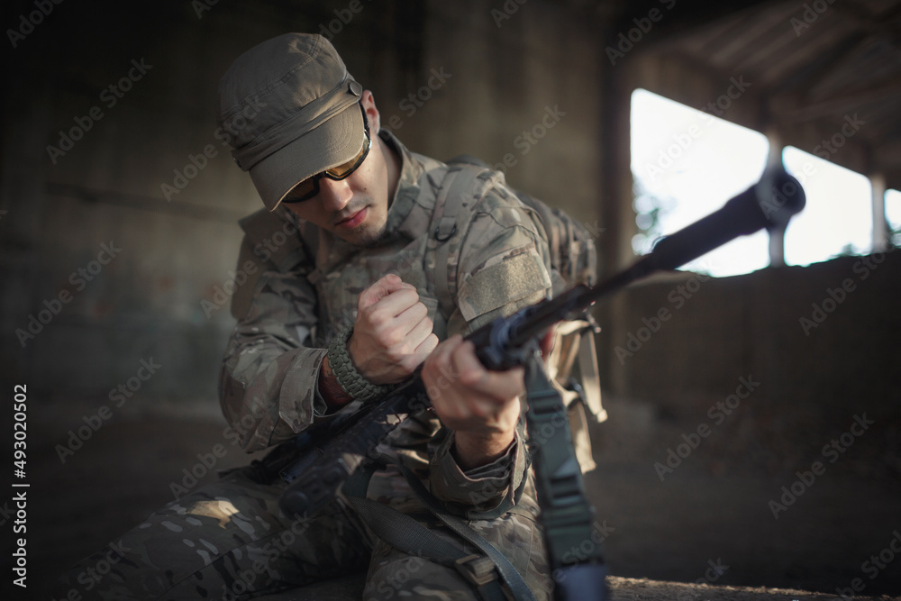 Fototapeta premium a Ukrainian soldier in a gray camouflage uniform wearing a cap and tactical black goggles sits on a concrete floor inside a large destroyed building and reloads a machine gun