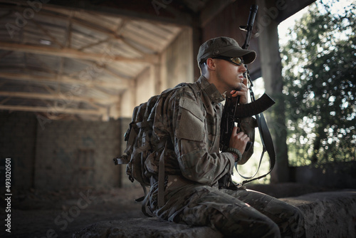 a soldier with a machine gun in his hands in a gray camouflage uniform with a large backpack on his back, wearing a cap and tactical glasses sits on a concrete floor inside a large destroyed building