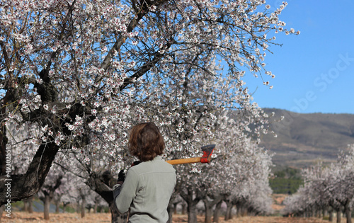 Young farmer with an axe on his shoulder in a field of flowering almond trees