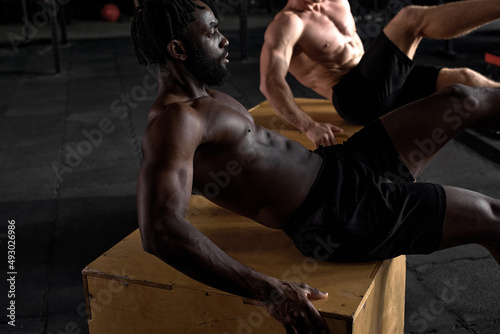 side view on fit muscular two men sitting on cross fit plyobox training legs and body, raising leg up leaning body on wooden box. focus on black african strong male concentrated on workout in gym