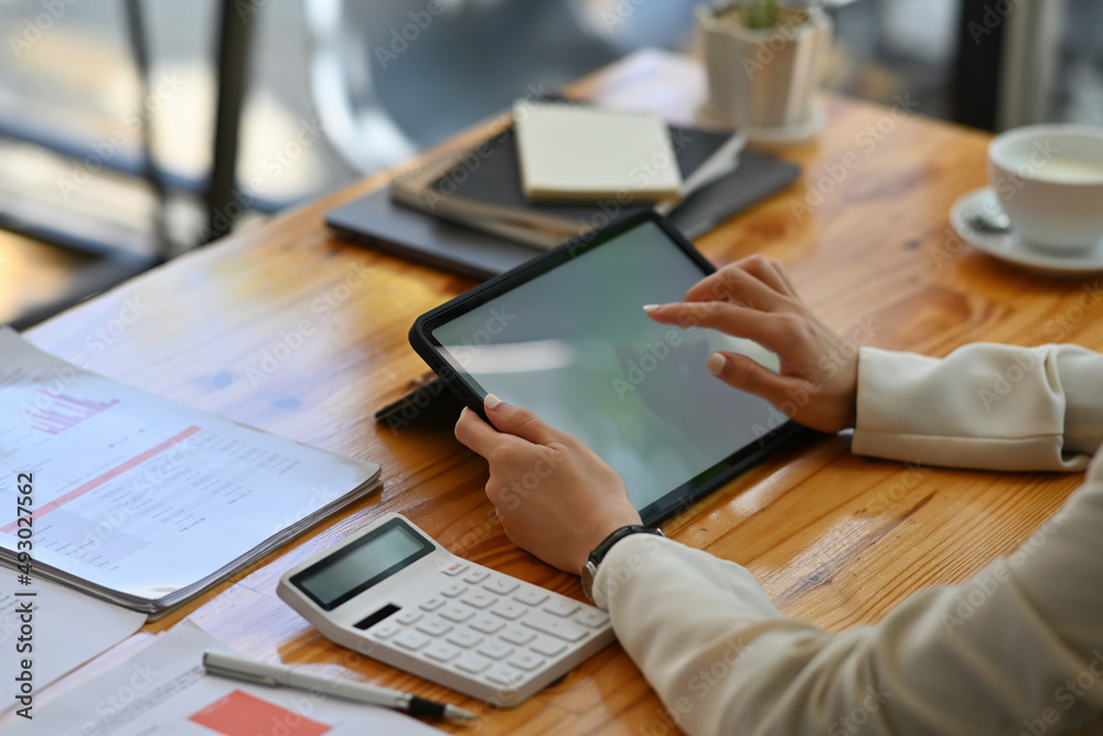 Cropped view young woman sitting in the office using a blank white screen tablet and  and a calculator working on data, charts and documents on the table, for business, finance and technology concept.