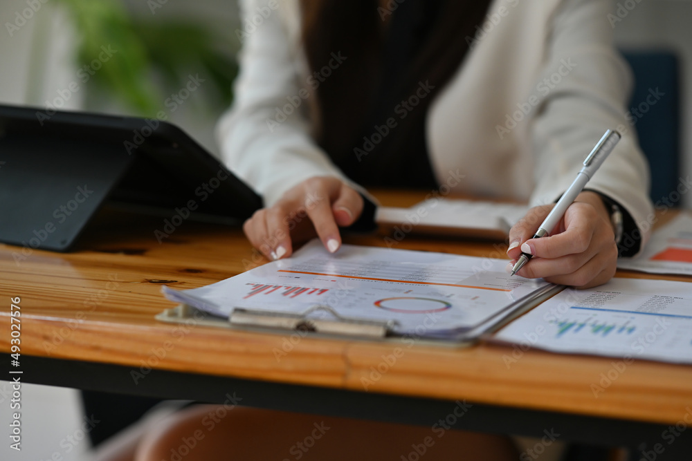 Cropped view young woman sitting in the office using a tablet and holding a pen working on data, charts and documents on the table, for business, finance and technology concept. Cropped view young woman sitting in the office using a tablet and holding a pen working on data, charts and documents on the table, for business, finance and technology concept.