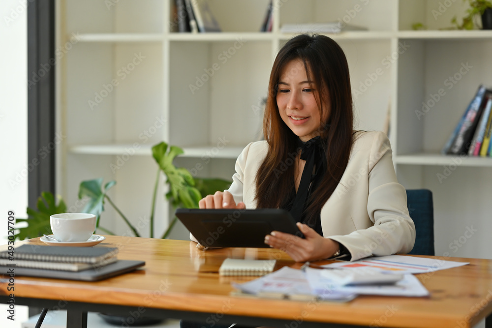 A portrait of a young pretty Asian woman sitting in the office using a tablet, calculator, working on data, charts and documents on the table, for business, finance and technology concept. A portrait of a young pretty Asian woman sitting in the office using a tablet, calculator, working on data, charts and documents on the table, for business, finance and technology concept.
