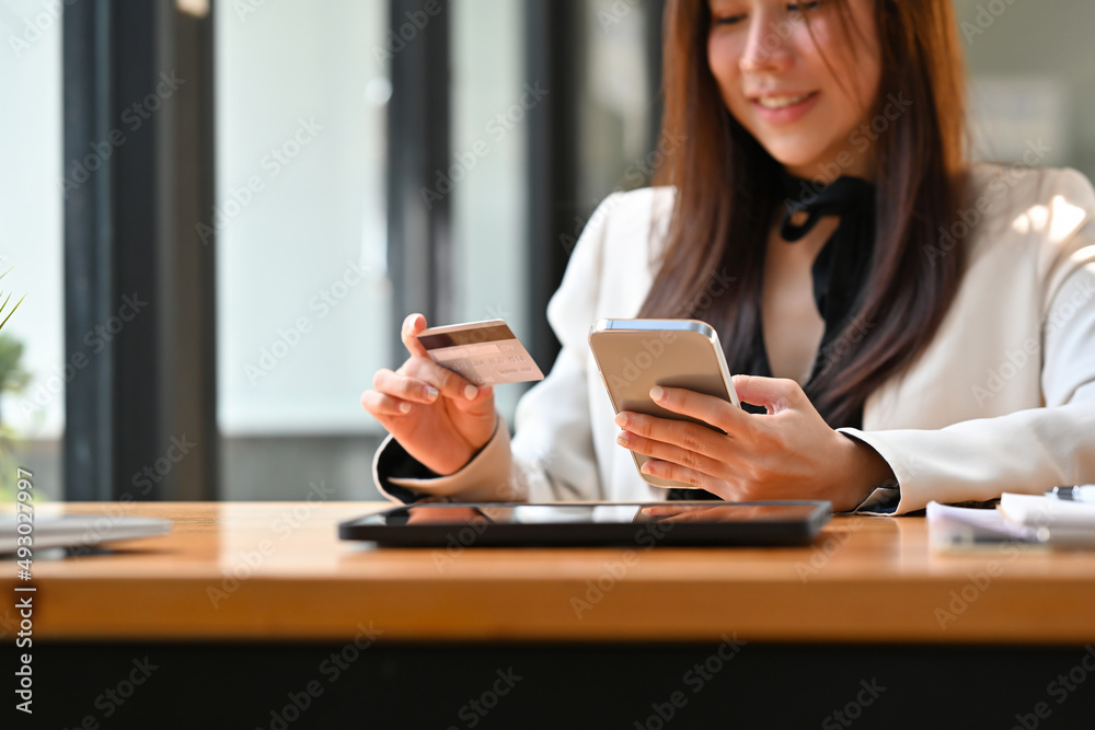 Cropped view of a pretty smiling Asian woman wearing a suit sitting in the office holding a credit card and a smart phone, for finance and technology concept. Cropped view of a pretty smiling Asian woman wearing a suit sitting in the office holding a credit card and a smart phone, for finance and technology concept.