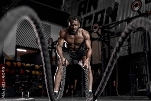 Portrait of strong young black man exercising with battle ropes during workout in modern gym, alone. indoors. active male having intense cross fit training, in dark sports club. fitness concept