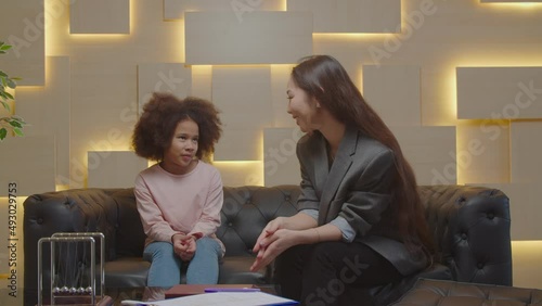 Caring positive pretty Asian female psychologist talking with upset adorable preadolescent African girl, helping to overcome mental problems and improve social skills during psychotherapy session. photo