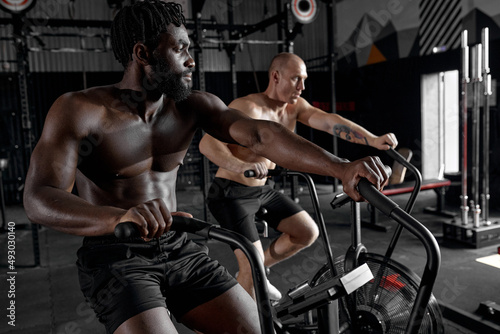 Fit young diverse men using exercise bike at modern gym. Fitness male using bike for cardio workout at cross training club, side view on concentrated sporty male, focused on cardio workout
