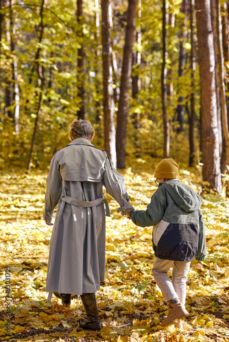 View from back on caucasian woman walking with child boy son, outdoors in park. short-haired blonde lady and boy holding hands together, have conversation, talking. close relationships