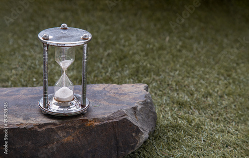 Silver hourglass or Sandglass on Huge stone over grass background. Hope and Value of time concept, Copy space, Selective focus. © num